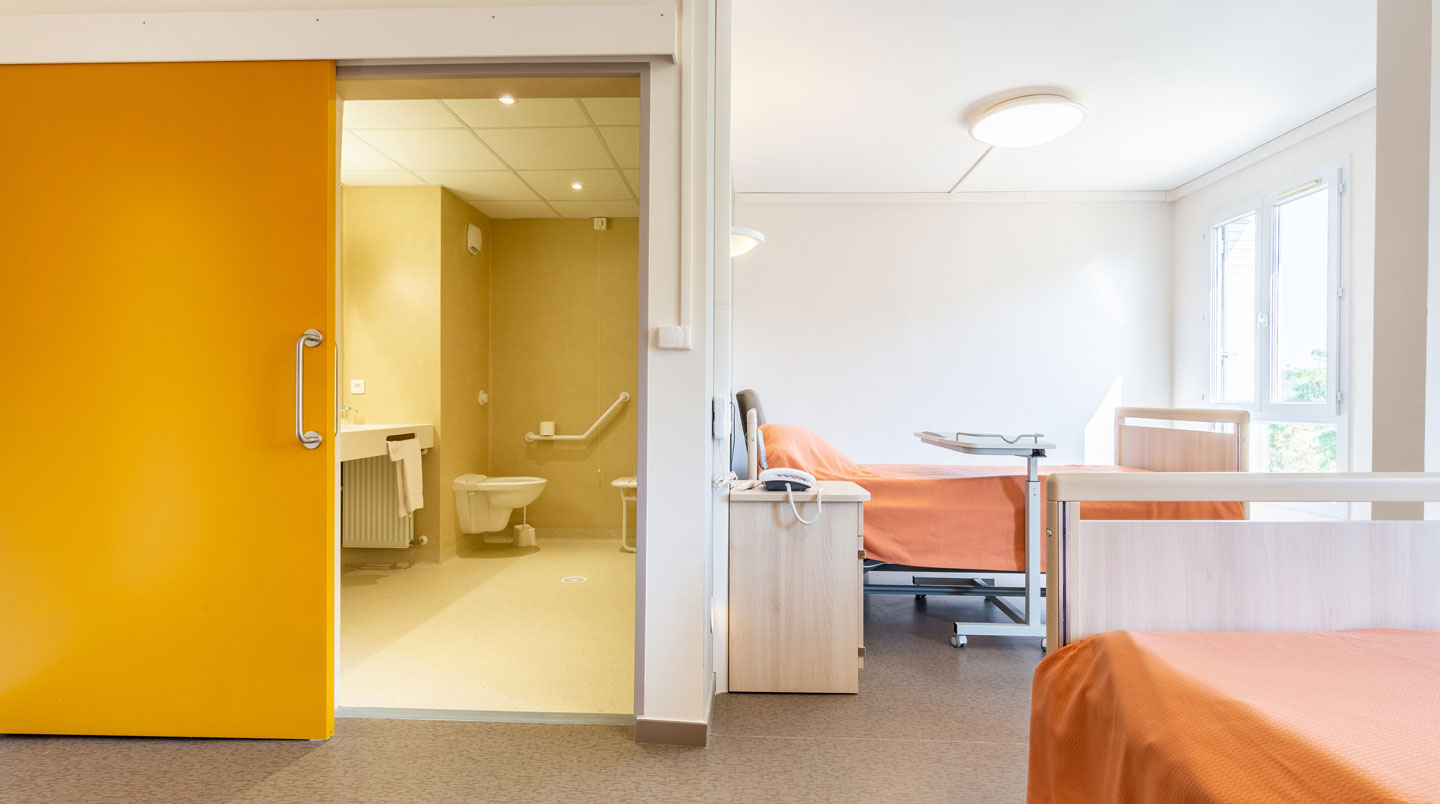 Vue grand angle d'un salon commun contemporain d'EHPAD avec de larges baies vitrées, des fauteuils confortables et une décoration sobre et lumineuse