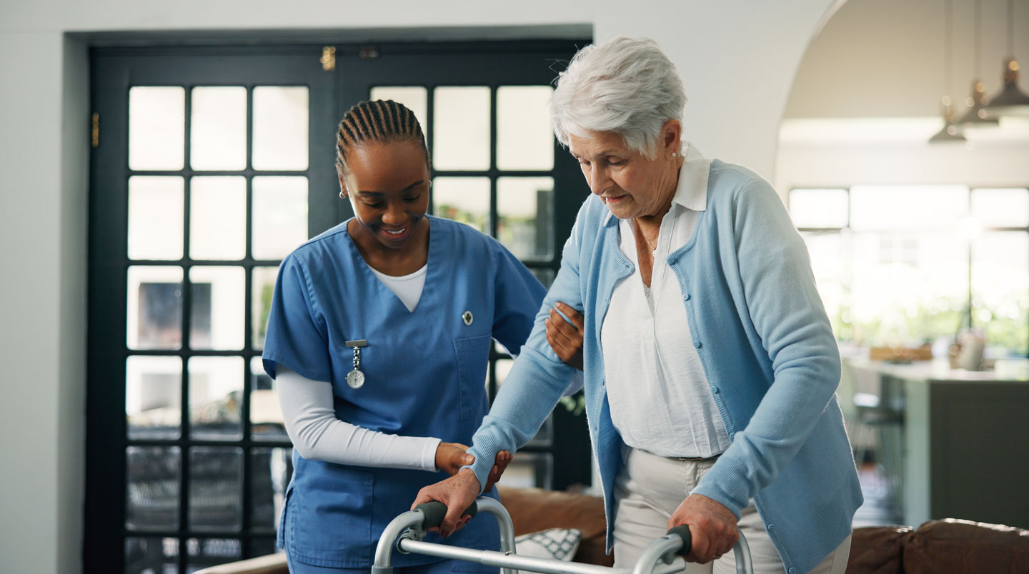 Une aide à domicile professionnelle accompagne un senior dans son salon pour l'aider à se déplacer en toute sécurité avec un déambulateur.